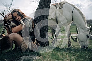 Woman rests on grass in image of warrior amazon with sword near grazing horse