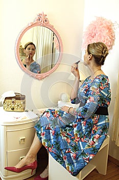Woman in a retro dress and heels looking at reflection at vanity preparing to put on lipstick