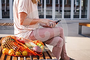Woman with reusable mesh bag sitting on bench and using smart phone in city