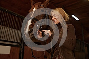 Woman rider putting bridle horsey muzzle while standing in stable