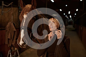 Woman rider putting helmet on head standing front of harnessed horse