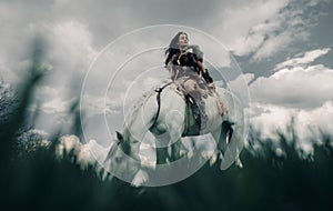 Woman rides on horseback in image of warrior amazon on grass and sky background