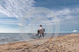 Woman is riding on a beautiful brown horse along a beach near the sparkling ocean