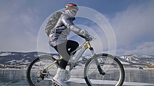 Woman is riding bicycle on the ice. The girl is dressed in a silvery down jacket, cycling backpack and helmet. Ice of