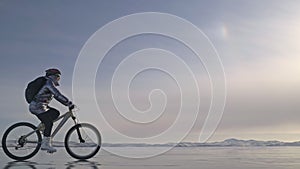 Woman is riding bicycle on the ice. The girl is dressed in a silvery down jacket, cycling backpack and helmet. Ice of