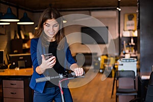 Woman riding electric scooter and using mobile phone at work in office.