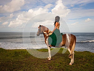 Woman riding horse near the ocean. Outdoor activities. Asia woman wearing long green dress. Traveling concept. Cloudy sky. View