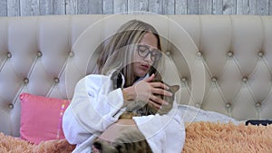 A woman in a robe is lying in bed with a cat in her hands and stroking the cat