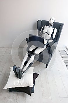 Woman robot working with laptop while sitting in cozy armchair putting her legs on pillow