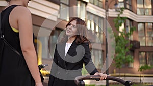 A woman rocks a baby stroller and talks to her friend while standing outdoors on a summer day in the courtyard of an