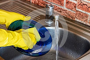 Woman in rubber yellow gloves wash dishes and plates with sponge in the metal sink at the kitchen.