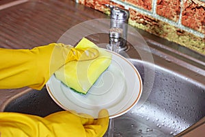 Woman in rubber yellow gloves wash dishes and plates with sponge in the metal sink at the kitchen.
