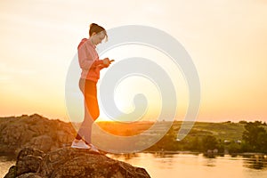 Woman Runner Resting after Workout, Using Smartphone and Listening to Music at Sunset on the Rock. Sports Concept