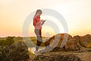 Woman Runner Resting after Workout, Using Smartphone and Listening to Music at Sunset on the Rock. Sports Concept