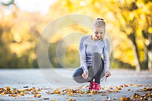Woman runner tying shoelaces before jogging in autumn tree alley park. Sports female autumn outfit leggings and thermal underwear