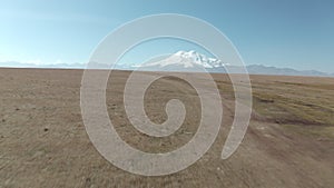 Woman running in field on snowy Elbrus peak landscape. Aerial view girl white t-shirt denim jumpsuit on highland field