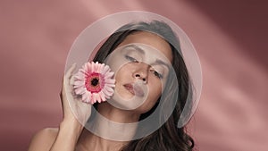 A woman runs a gerbera flower across her perfect skin. Portrait of a woman with a gerbera flower in the studio on a pink