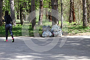 Woman runs into park next to plastic bags full of waste. Plastic pollution on environment. Woman does sport in the forest.