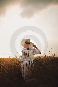 Woman in rustic dress and hat enjoying peaceful sunset in wildflowers and herbs in summer meadow. Atmospheric authentic moment.