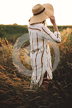 Woman in rustic dress and hat enjoying peaceful sunset in wildflowers and herbs in summer meadow. Atmospheric authentic moment.