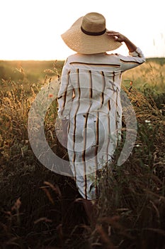 Woman in rustic dress and hat enjoying peaceful sunset in wildflowers and herbs in summer meadow. Atmospheric authentic moment.