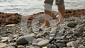 A woman's bare feet walk on a rocky sea beach. Dried seaweed, plastic trash, and water in the background. Close-up.