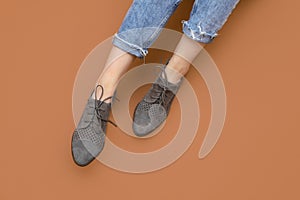 Woman`s Caucasian legs in blue twisted jeans and gray suede English brogue Oxfords shoes isolated on Brown beige background.