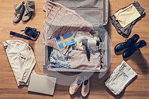 Woman`s clothes, laptop, camer and flag of Sweden lying on the parquet floor near and in the open suitcase. Travel photo