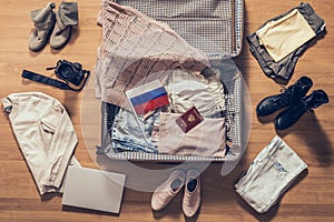 Woman`s clothes, laptop, camera, russian pasport and flag lying on the parquet floor near and in the open suitcase. Travel photo