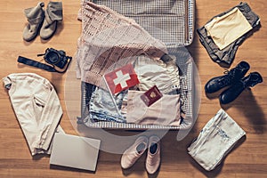 Woman`s clothes, laptop, camera, russian passport and flag of Switzerland lying on the parquet floor near and in the open suitcas