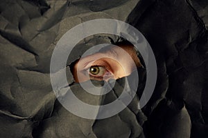 Woman`s eye in hole in black crumpled paper background, minimalist concept photography for blog or poster