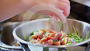 Woman's hand adding salt in metal bowl with vegetable salad and mixing it with steel spoon, closeup with slow motion