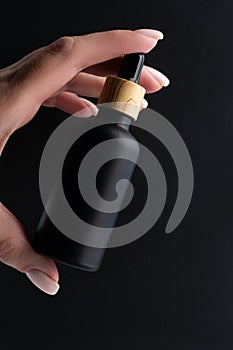Woman& x27;s hand with a black frosted glass bottle of beauty oil on a black background. Cosmetic products concept. Copy
