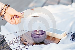 Woman`s hand in bracelet with purple long nails lights candle with match, next to deck of cards and lavender flowers