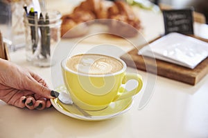 Womanï¿½s hand and cappuccino coffee in yellow cup and saucer