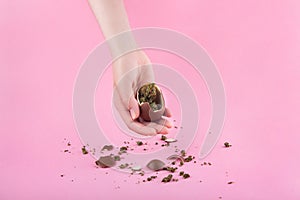 In a woman's hand is a chocolate egg with dry buds of medical marijuana inside. Pieces of chocolate and buds are scattered