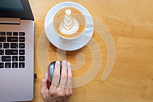 Woman`s hand clicking on wireless mouse with laptop and cup of coffee besides on wooden table.Top view.