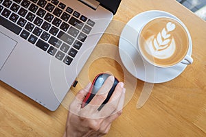 Woman`s hand clicking on wireless mouse with laptop and cup of coffee besides on wooden table.Top view.