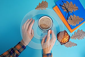 Woman`s hand with cup of coffee, books and cake on a autumn blue background