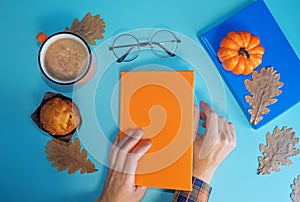 Woman`s hand with cup of coffee, books and cake on a autumn blue background