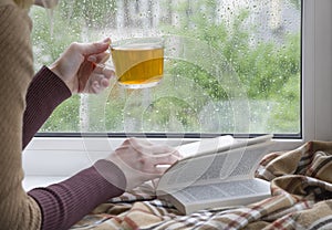A woman& x27;s hand with a cup of tea. An open book and a soft blanket.