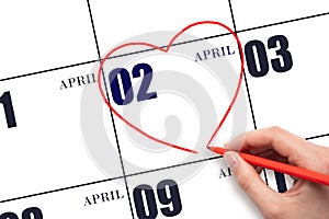 A woman's hand drawing a red heart shape on the calendar date of 2 April. Heart as a symbol of love.