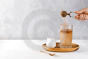 Woman`s hand dusting cocoa into vegan iced coffee in a tall glass on a wooden tray