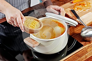 Woman`s hand hold a wood bowl with raw noodles to put it into the boiling bouillon. Opened white saucepan on the black stove.