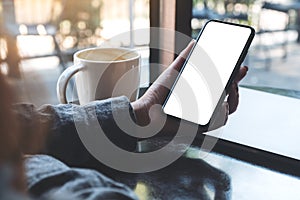 A woman`s hand holding black mobile phone with blank white desktop screen with coffee cup and laptop on table in cafe