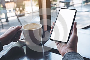 A woman`s hand holding black mobile phone with blank white desktop screen with laptop and coffee cup on table in caf