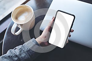 A woman`s hand holding black mobile phone with blank white desktop screen with laptop and coffee cup on table in cafe