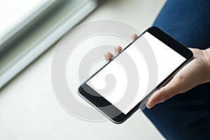 Woman`s hand holding black mobile phone with blank white screen on thigh with white tile floor and sliding door background