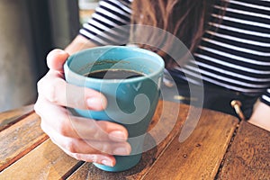 A woman`s hand holding blue mug of hot coffee on vintage wooden table in cafe