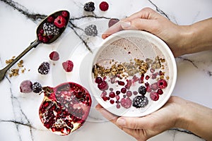 Woman`s hand holding a bowl of yoghurt with cereals and forest fruits. Pomegranate on the table besides.